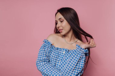 Mavi kareli omuzlu bir kadın pembe bir arka plana karşı poz veriyor, bir eliyle nazikçe saçlarını tutuyor ve sakin bir ifadeyle aşağıya bakıyor..