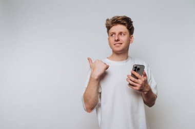 Beyaz tişörtlü genç adam elinde akıllı bir telefon tutuyor ve arkasını işaret ediyor..