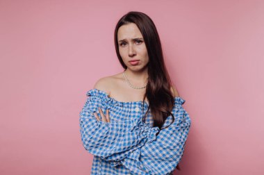 Mavi omuzlu, uzun kahverengi saçlı bir kadın pembe bir arka plana karşı duruyor, kollarını kavuşturmuş üzgün görünüyor..