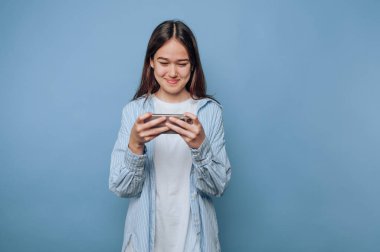 Mavi arka planda akıllı telefon kullanan genç bir kadın gülümsüyor..