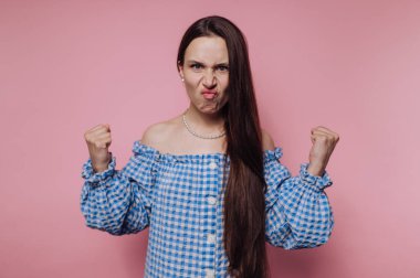 Mavi kareli omuzlu bir kadın pembe bir arka plana karşı yumruklarını sıkmış öfkesini ifade ediyor..