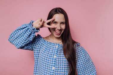 Mavi kareli bluzlu neşeli bir kadın pembe arka planda şakacı poz veriyor..