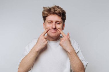 Beyaz gömlekli genç adam gülümsüyor ve yanaklarını çekiyor..