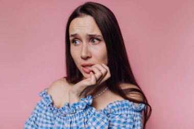 Uzun siyah saçlı, pembe arka planda tırnaklarını yiyen endişeli bir kadın..