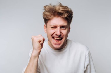 Açık kahverengi saçlı genç bir adam heyecan içinde yumruğunu sıkar, düz bir arkaplanda beyaz bir gömlek giyer..