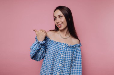 Mavi omuzlu genç bir kadın pembe arka planda arkasını işaret ediyor..