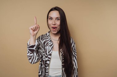 Uzun saçlı, şaşkın bir kadın işaret parmağını kaldırır ve bej bir arka plana zebra desenli bir gömlek giyer..
