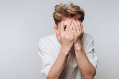 Beyaz gömlekli bir adam iki eliyle yüzünü kapatır ve parmaklarından içeri bakar..