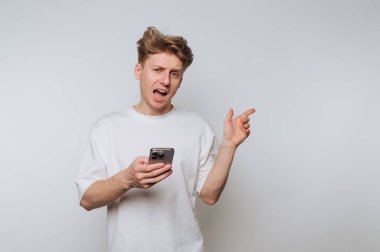Beyaz tişörtlü genç adam elinde akıllı bir telefon tutuyor, işaret ediyor ve şaşırmış görünüyor..