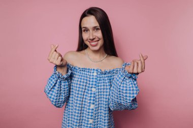 Mavi omuzlu gülümseyen kadın pembe arka planda parmaklarıyla kalp hareketleri yapıyor..
