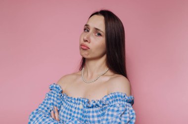 Üzerinde inci kolyesi olan mavi omuzlu bir kadın pembe arka planda duruyor, kolları bağlı dalgın dalgın bakıyor..