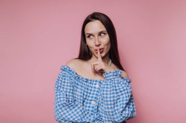 Mavi kareli omuzlu bir kadın pembe arka planda poz veriyor, parmaklarını şakaya vurarak dudaklarına dokunuyor..