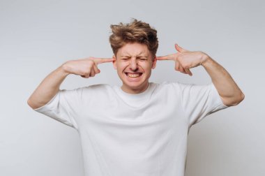 Beyaz tişörtlü genç bir adam kulaklarını parmakları, gözleri kapalı bir şekilde kapatır, hayal kırıklığını ya da sıkıntıyı arka planda gösterir..