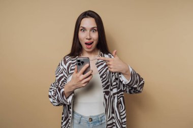 Şaşırmış bir kadın bej arka planda akıllı telefonunu işaret ediyor..