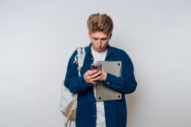 Sırt çantalı genç bir adam dizüstü bilgisayar ve akıllı telefon tutuyor, şaşırmış görünüyor..