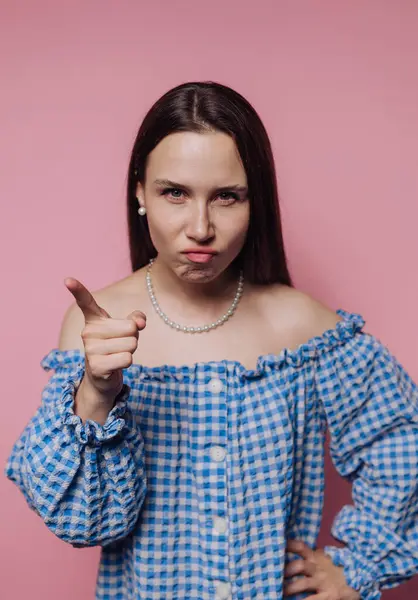 Mavi kareli elbiseli kadın pembe arka planda ciddi bir ifadeyi işaret ediyor..