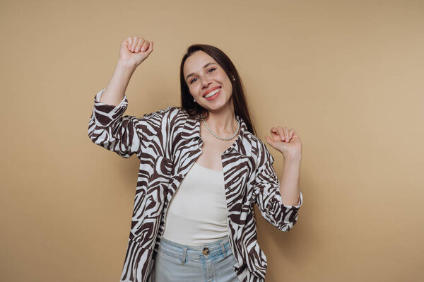 A joyful young woman with long dark hair wearing a stylish zebra-patterned shirt and light blue jeans. She is smiling and dancing against a warm beige background, exuding happiness and confidence.