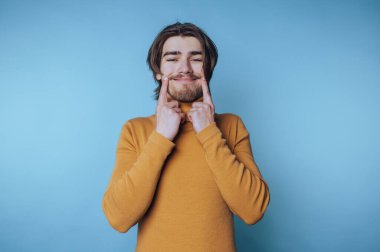 Balıkçı yakalı genç bir adamın portresi parmaklarıyla gülümseyerek mavi arka planda pozitiflik yayıyor.