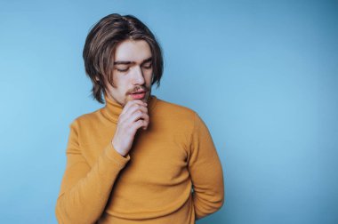 Balıkçı yaka giyen genç bir adamın portresi düşünceli bir şekilde mavi arka planda dalgın dalgın duruyor.