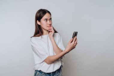 Genç bir kadının portresi cep telefonunu tutarken düşünceli görünüyor, arka planda önemli bir şeyi yansıtıyor.