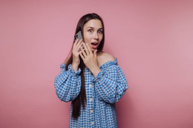 Şaşırmış genç bir kadının portresi mavi omuzsuz bir bluz giyerken sevinçle telefona tepki veriyor, pembe arka planda keyif ve heyecanı somutlaştırıyor.