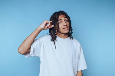 Örgülü saçlı genç bir adamın portresi, mavi arka planda düşünceli bir şekilde, parmağını şakağına uzatan akıllı bir ifadeyi gösteriyor.