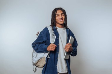 Rastalı genç bir adamın portresi kitaplar ve sırt çantasıyla poz veriyor, hafif arka planda eğitimi simgeliyor.