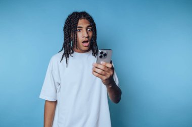 Genç adamın portresi, mavi arka planda açıkça şok olan akıllı telefonlarına bakıyor.