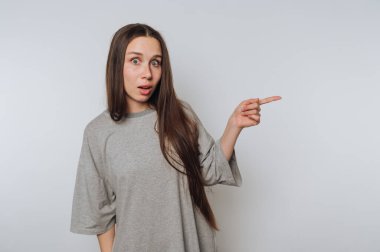 Uzun saçlı, şaşırmış genç bir kadının portresi açık arkaplanda kamera dışı bir şeye işaret ediyor.