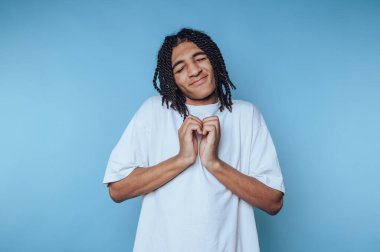 Saçları örgülü genç bir adamın portresi neşe ve sevgiyi ifade eder, mavi arka planda vücut dilinde duygularını gösterir.