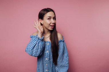 Uzun saçlı genç bir kadının portresi pembe arka planda bir merak göstergesi olarak elini kulağına kaldırır.