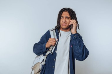 Genç bir adamın portresi açık arkaplanda sırt çantası tutarken telefonuyla konuşuyor.