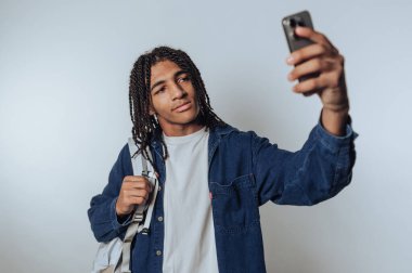 Genç bir adamın portresi, akıllı telefonu ve sırt çantasıyla özçekim çekerken hafif arka planda kendine güvenini yayar.