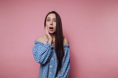 Mavi kareli kıyafetli genç bir kadının portresi, pembe arka planda eli yanağında bir şok gösteriyor.