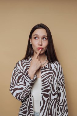 Düşünceli genç bir kadının portresi, sakin sarı arka planda modaya uygun zebra desenli bir gömlek giymiş, dudaklarında bir parmak ile poz veriyor.