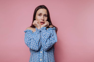 Kareli gömlekli genç bir kadının portresi pembe arka planda endişeli bir görünüm sergiliyor.