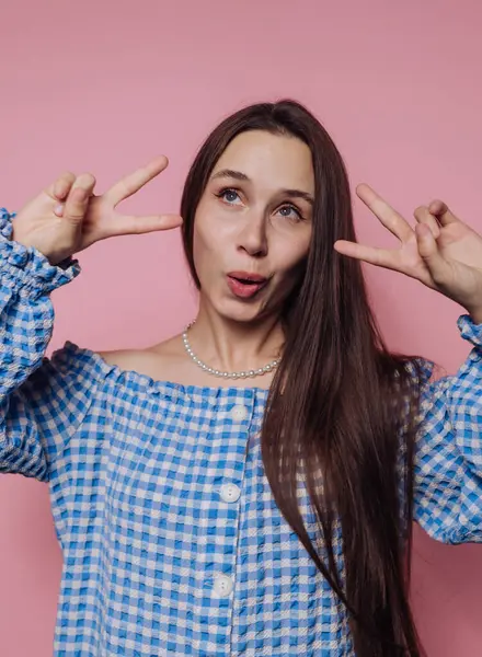 Genç bir kadının portresi pembe arka plandaki barış işaretinde parmaklarıyla poz veriyor.