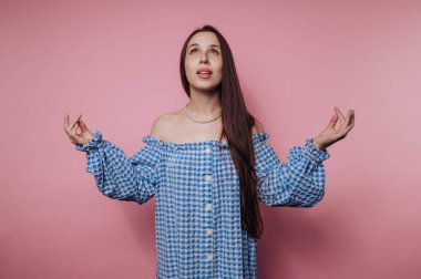 Bir kadın ellerini kaldırmış, sanki meditasyondaymış gibi, mavi ve beyaz bir gingham elbiseyle zarif bir şekilde durur. Pembe bir arka plan atmosferi canlandırırken nazikçe gülümsüyor..