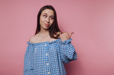 Pembe bir duvarın önünde duran genç bir kadın mavi kareli, omuzsuz bir gömlek giyiyor. Şakacı bir tavır sergiliyor, başparmağıyla işaret ediyor ve gülümsüyor..