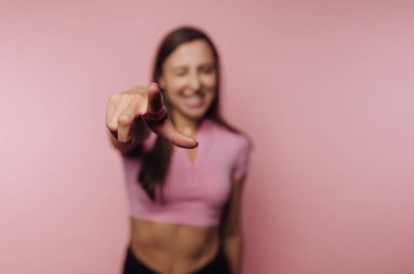 Pembe bluzlu genç bir kadın neşeli bir ifadeyle ayakta duruyor, neşeli bir şekilde parmağını pembe bir arka plana doğru gösteriyor, neşeli ve kaygısız bir hava yayıyor..