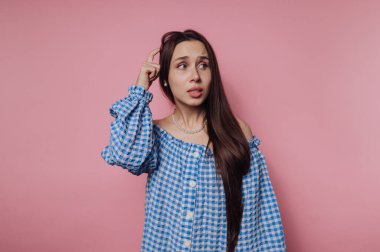 Mavi ve beyaz kareli bluz giymiş genç bir kadın pembe bir zemine karşı duruyor. Uzun saçları var ve kafası karışmış bir ifadeyle bir soruyu düşünüyor gibi görünüyor..