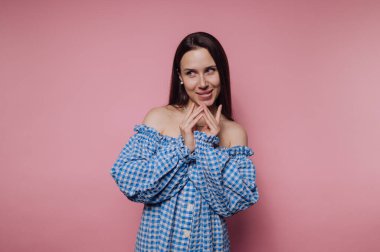 Bir kadın neşeli bir ifadeyle ayakta duruyor, elleri birbirine kenetlenmiş, canlı pembe bir arka plana karşı. Mavi ve beyaz, omuzları açık bir elbise giyiyor, neşeli tavırlarını sergiliyor..