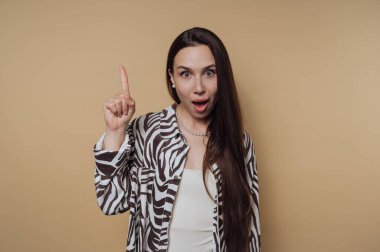 Bir kadın bej bir arka fonda duruyor, işaret parmağını oynak bir şekilde kaldırırken şaşkın bir ifade takınıyor, desenli bir ceket giymiş..