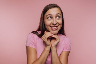 Pembe tişörtlü genç bir kadın, büyüleyici bir gülümsemeyle, elleri yüzüne yakın, yumuşak pembe bir arka planla stüdyoda heyecan yayıyor..