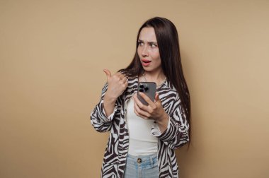 Genç bir kadın bej bir arka fonun önünde duruyor, bir elinde akıllı bir telefon tutarken şaşkınlığını dile getiriyor ve diğer elinde de başparmağını kaldırıyor. Şık bir gömlek ve rahat bir kot pantolon giyiyor..