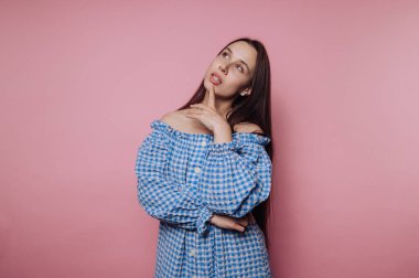 Omuzları açık mavi bir elbise giyen bir kadın pembe bir arkaplanın önünde duruyor, stüdyo ortamında düşünce ve tarzın bir yansımasını canlandırıyor..