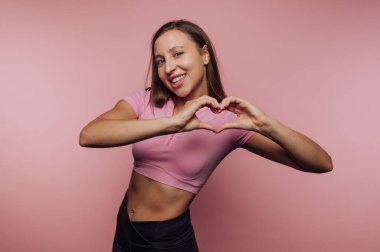 Gülümseyen genç bir kadın elleri ile canlı pembe bir arka plan oluşturur. Neşeli bir şekilde poz verirken üstündekini çıkarıp neşe ve pozitiflik saçıyor..