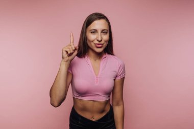 Bir kadın pembe bir zemine karşı duruyor, gülümsüyor ve şakasına parmağını kaldırıyor. Günlük kıyafetiyle kendine güveni ve ulaşılabilirliği yayıyor, dostane bir ortam yaratıyor..