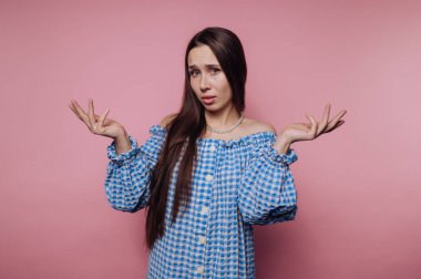 Uzun saçlı genç bir kadın mavi kareli bir elbise giyerek elleriyle karışık bir jest yapıyor. Arkaplan yumuşak pembe, neşeli bir atmosfer yaratıyor..