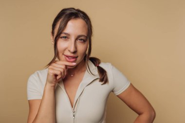 Bir kadın, neşeli bir sırıtışla, rahat bir şekilde poz verir. Bir eli çenesinde duruyor, ifadesini arttıran yumuşak, nötr bir zemin üzerinde duruyor..